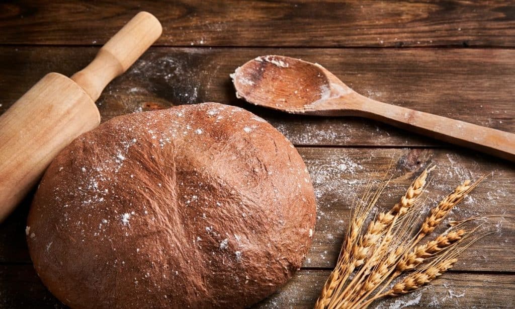 A round bread with flour sprinkled, along with a rolling pin and a wooden spoon. 
