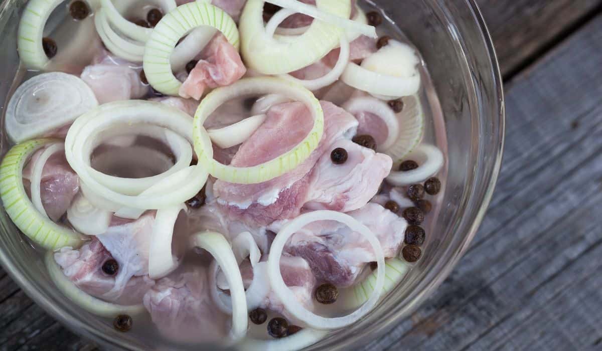 A clear bowl on a dark wood surface with meat, clear brine, onion slices, and peppercorns.